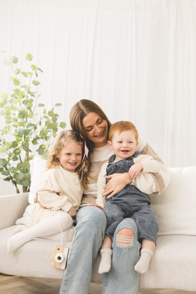 Mum hugging her one year old baby and toddler daughter with big smiles on a sofa with a plant behind